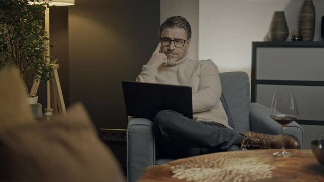 Middle aged man sitting in comfort in living room working on laptop computer in home office. Businessman managing business online. Portrait of confident older male, happy, smiling, thinking.  .