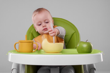 Cute little baby eating healthy food in high chair on gray background