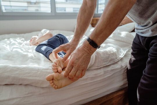 Grandpa playing with his little grandson tickling his barefoot feet. Joyful toddler boy having fun playing in bedroom with dad. Focus on hands.