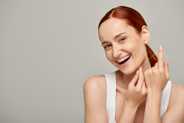 excited redhead model holding dental floss and smiling on grey background,  promoting oral hygiene