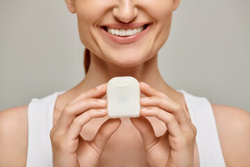 cropped view of positive redhead woman holding dental floss case and smiling on grey background
