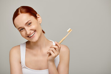 happy woman with red hair holding bamboo toothbrush with toothpaste on grey backdrop, dental
