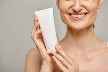 cropped young and happy woman in 30s holding tube with body lotion on grey background, skincare
