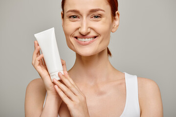 Redhead young and happy woman in 30s holding tube with body lotion on grey background, skincare