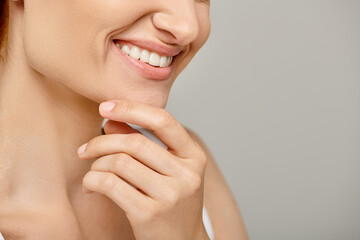 cropped view of woman with healthy smile touching radiant skin on grey background, close up