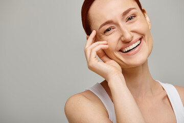 cheerful and redhead woman in white tank top looking at camera on grey background, healthy smile