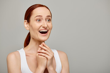 astonished and redhead woman in white tank top with healthy smile on a grey background, laughter