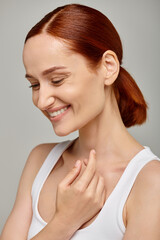 cheerful redhead woman in white tank top exuding joyful and healthy smile on grey background