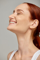 happy redhead woman with closed eyes exuding joyful and healthy smile on grey background - 761631893