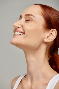 happy redhead woman with closed eyes exuding joyful and healthy smile on grey background
