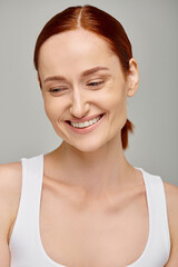 happy redhead woman in tank top exuding joyful and healthy smile on grey background, looking down