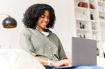 With a warm smile, the African-American woman enjoys her time on the laptop, perfectly blending productivity with the comfort of her home environment