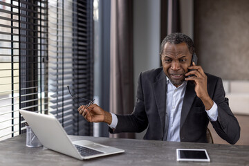 Furious diverse male in formalwear holding eyeglasses while having unpleasant phone conversation. Angry distant worker hearing bad breaking news while sitting by desktop with portable computer