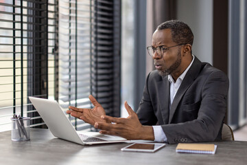 Clueless business person spreading hands to computer while working by table with tablet and notebook. Mature man in jacket trying solving company processes and feeling desperate about new tasks