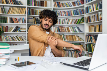 Young tired guy stretching hand and shoulder while looking at notebook on desk with books and computer. Bored male making light exercise for reducing muscle tension before going back to work.