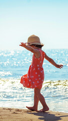 Happy little girl with raised arms walks along the beach in summer back to the camera. Baby in an orange dress with polka dots and a straw hat. Vertical banner - 761659861