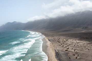 Aerial view of a wavy ocean and sandy dunes with some vegetation, with spectacular mountains on the background, Lanzarote, Canary Islands