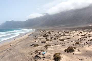 Aerial view of a wavy ocean and sandy dunes with people walking along the Famara beach, with cliffs on the background, Lanzarote, Canary Islands