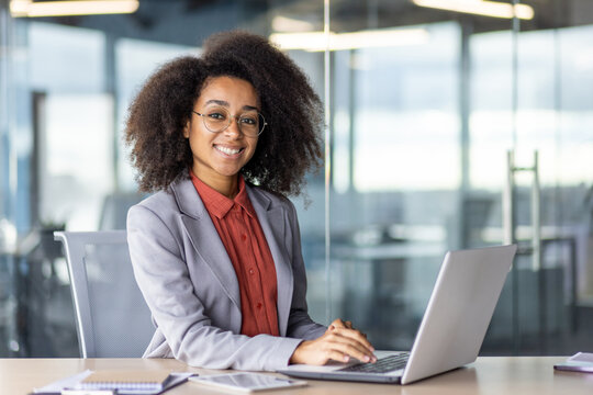 Attractive african american freelancer smiling at camera while keeping hands on portable computer keyboard. Successful interior designer enjoying productive busy day in modern shared coworking.