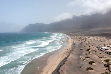 Aerial view of a wavy ocean and surfers along the Famara beach, with dunes and spectacular mountains on the background, Lanzarote, Canary Islands