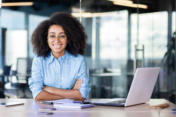 Successful black woman in blue shirt leaning with folded hands on table with laptop and daybook. Smiling lady looking at camera during working day as secretary in leading financial company.