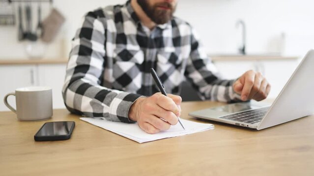 Close up of hand of businessman with pen making notes on draft of paper document being placed near laptop on wooden desk. Well-organised man remote employee starting business day in workplace at home.