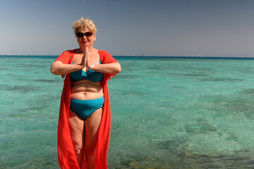 senior woman wearing in swimsuit and sunglasses and doing yoga and meditating on the sea beach with clear water in sunny day.