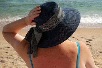 a woman in a swimsuit and a large red hat sits on the beach against the backdrop of the sea on a sunny summer day
