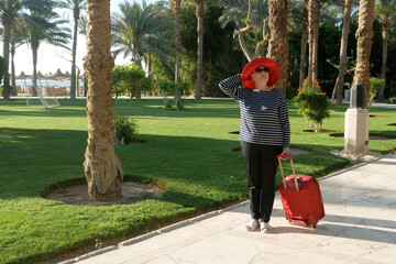 Middle aged blond woman wearing in red hat with a travel bag is settled in the hotel resort for a vacation, passing by alley with palm trees against the backdrop of the beach and sea