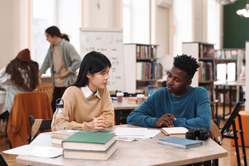 Portrait of young Asian woman and Black man sitting at table in class together during pair study