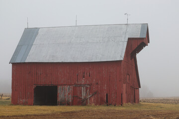 A stately, old, red barn slowly becoming engulfed by an evening fog.  - obrazy, fototapety, plakaty