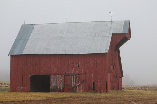A stately, old, red barn slowly becoming engulfed by an evening fog. 