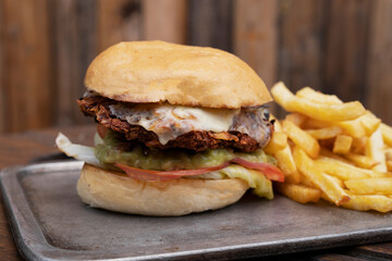 Fast food. Closeup view of a chicken burger with potato bread, cheese, lettuce, tomato, guacamole and french fries, in a metal dish on the wooden table.	
