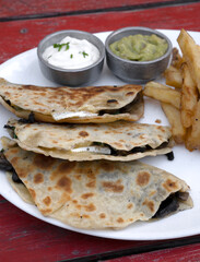 Mexican cuisine. Closeup view of quesadillas filled with cheese, french fries and dipping sauces, in a white dish on the restaurant wooden table	