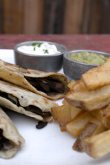 Mexican cuisine. Closeup view of quesadillas filled with cheese, french fries and dipping sauces, in a white dish on the restaurant wooden table	
