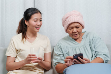 Elder in pink beanie looks at smartphone, young female holds empty glass, both seated indoors. Mature individual examines mobile phone screen, youthful companion with glass, sharing moment on couch.