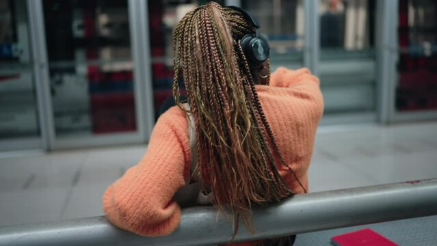 Young African woman listens to music in noise cancelation headphones suggesting immersion in private soundscape. Female technology user seated on bench at subway station, enjoy moment of leisure
