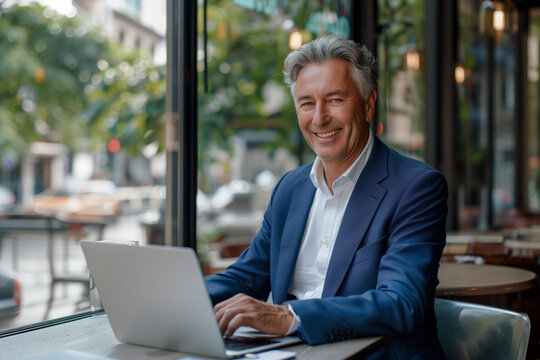 A high-resolution photo captures a middle-aged businessman, dressed in a blue suit and tie, smiling as he types on his laptop at a modern cafe with a cityscape view
