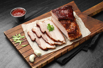 Pieces of baked pork belly served with sauce and parsley on black textured table