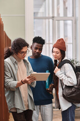 Vertical portrait of diverse group of three young students looking at tablet screen while standing in college library with backpacks