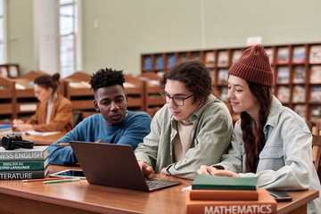 Multiethnic group of three young people using laptop together and doing research in college library...