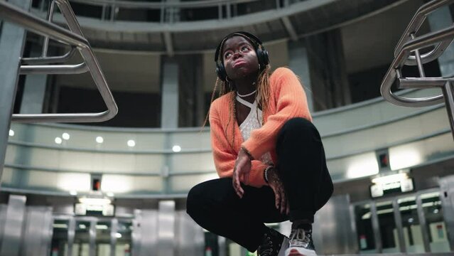 Contemplative female technology user enjoying music beat in headphones while sitting in urban subway station. African American young woman with vitiligo skin listen playlist in earbuds. Slow motion
