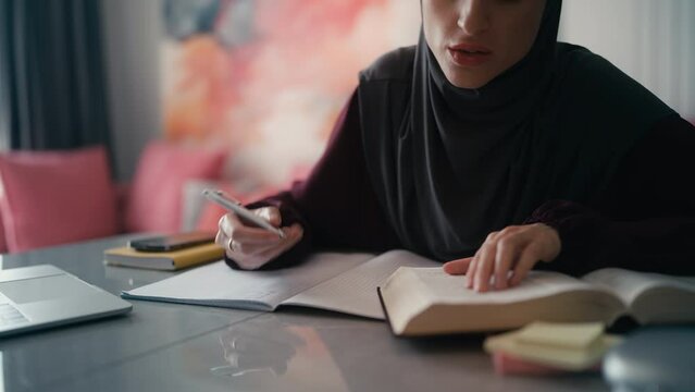 Female Arab student in hijab studying at home, reading textbook, writing notes