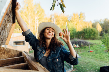A curly-haired woman in farm clothes beams as she takes an egg from a chicken coop. A woman shows...
