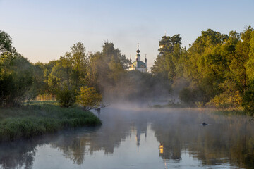 Church by the river, illuminated by the rays of the rising sun, mystical landscape with river and trees and fog
