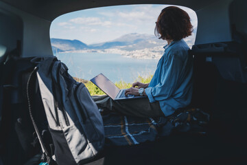 Young woman sitting in the open trunk of a car overlooking the sea and using a laptop, freelancer, remote work and summer vacation auto travel - 761733803