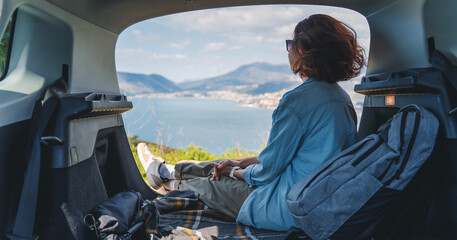 A young woman traveling by car sitting in the open trunk of a car admiring the view of the sea and...
