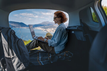 Young woman sitting in the open trunk of a car overlooking the sea and using a laptop, freelancer, remote work and summer vacation auto travel