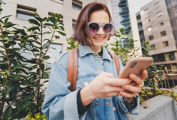 Young European woman in sunglasses with smartphone standing against the background of a modern residential area on a spring day - 761735476