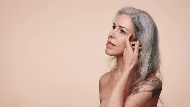 Confident mature woman with grey hair and floral tattoo looking thoughtfully touching skin. Ageing gracefully beauty concept.